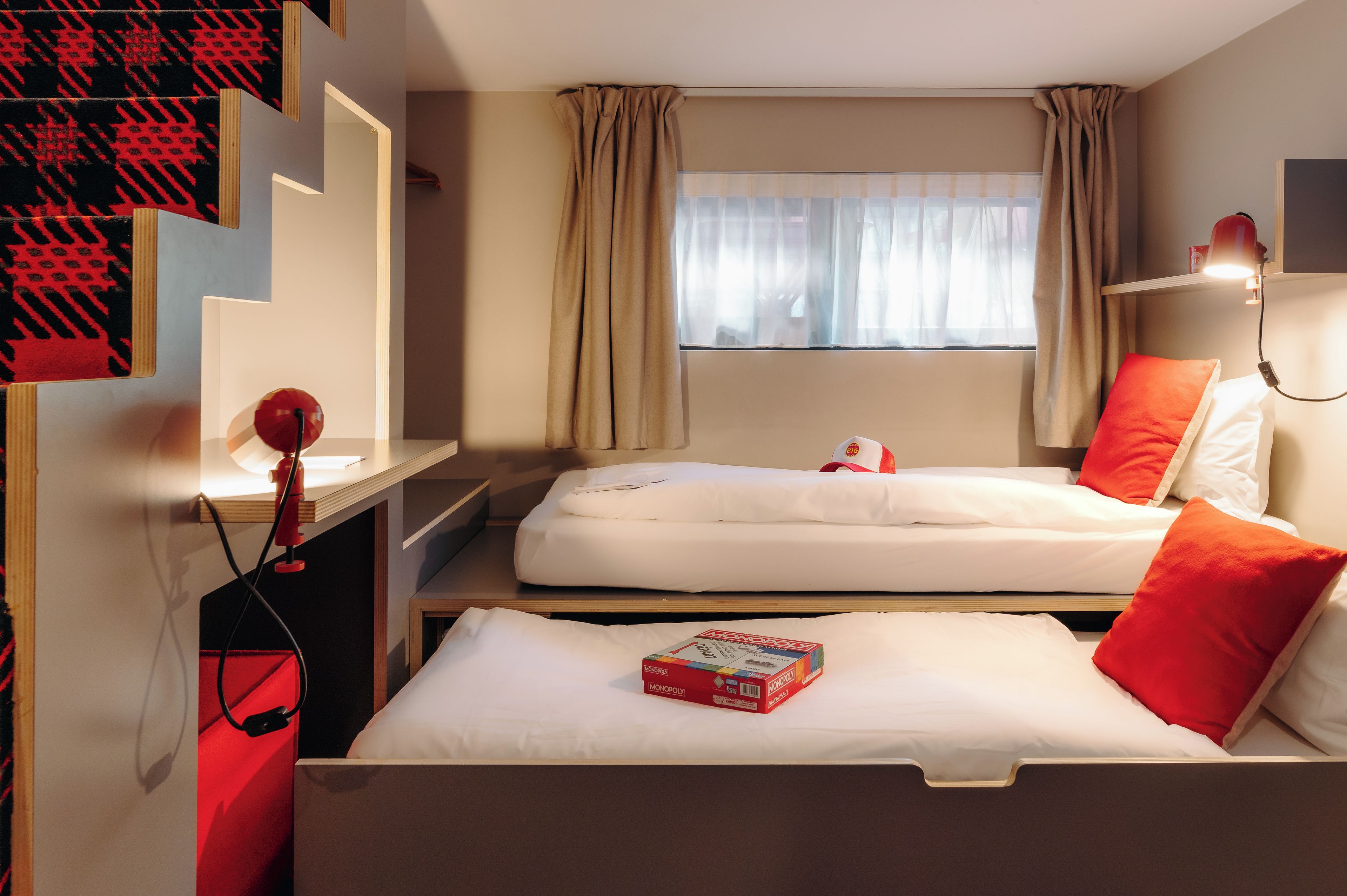 Ein Hotelzimmer mit zwei Betten, einem Schreibtisch, einem Stuhl, einem Fenster mit Vorhängen und einer roten Lampe.