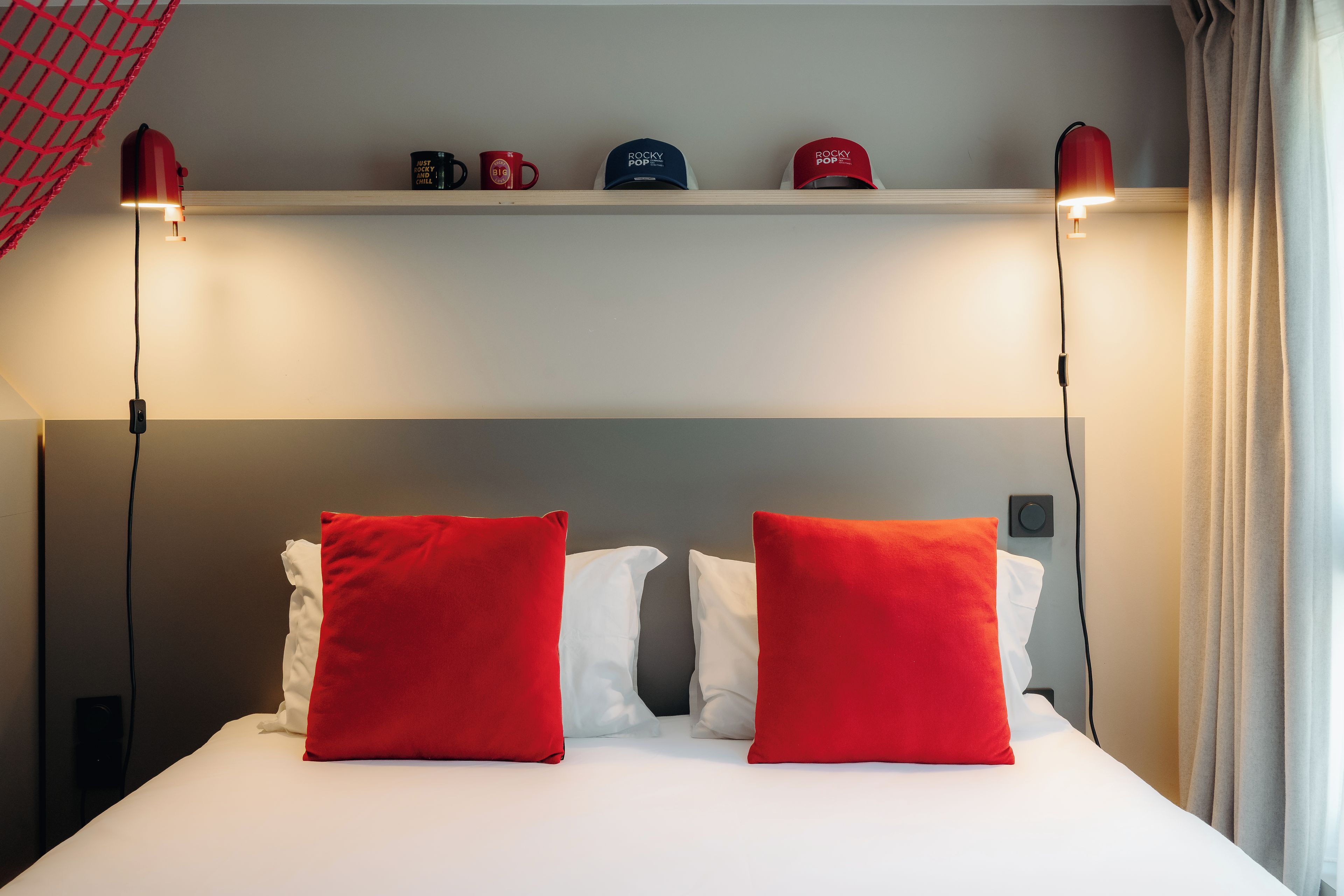 Una habitación de hotel con una cama, dos almohadas rojas, una repisa con objetos y dos lámparas de pared.