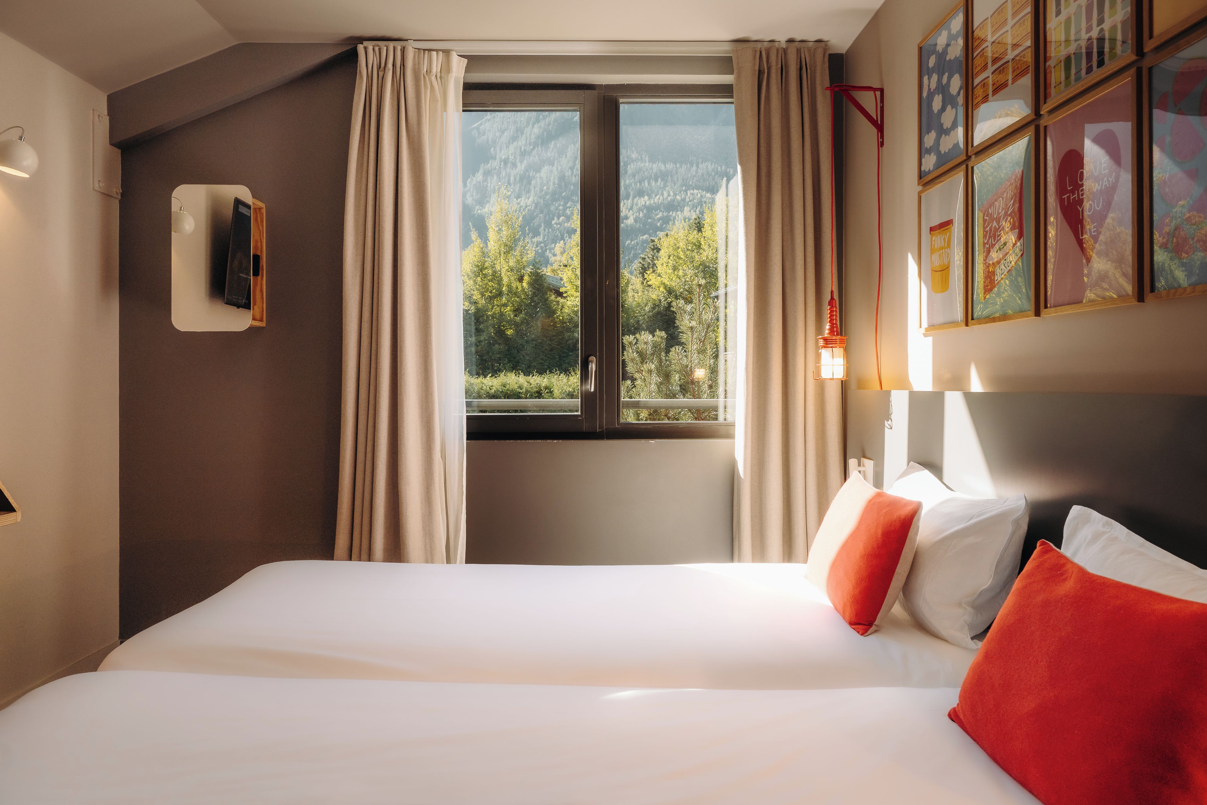 Habitación de hotel con una cama grande, una ventana con cortinas, una lámpara de pared e imágenes enmarcadas.