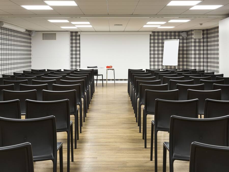 Un salón de conferencias con filas de sillas negras, una pantalla de proyección blanca y una pequeña mesa con un objeto rojo.
