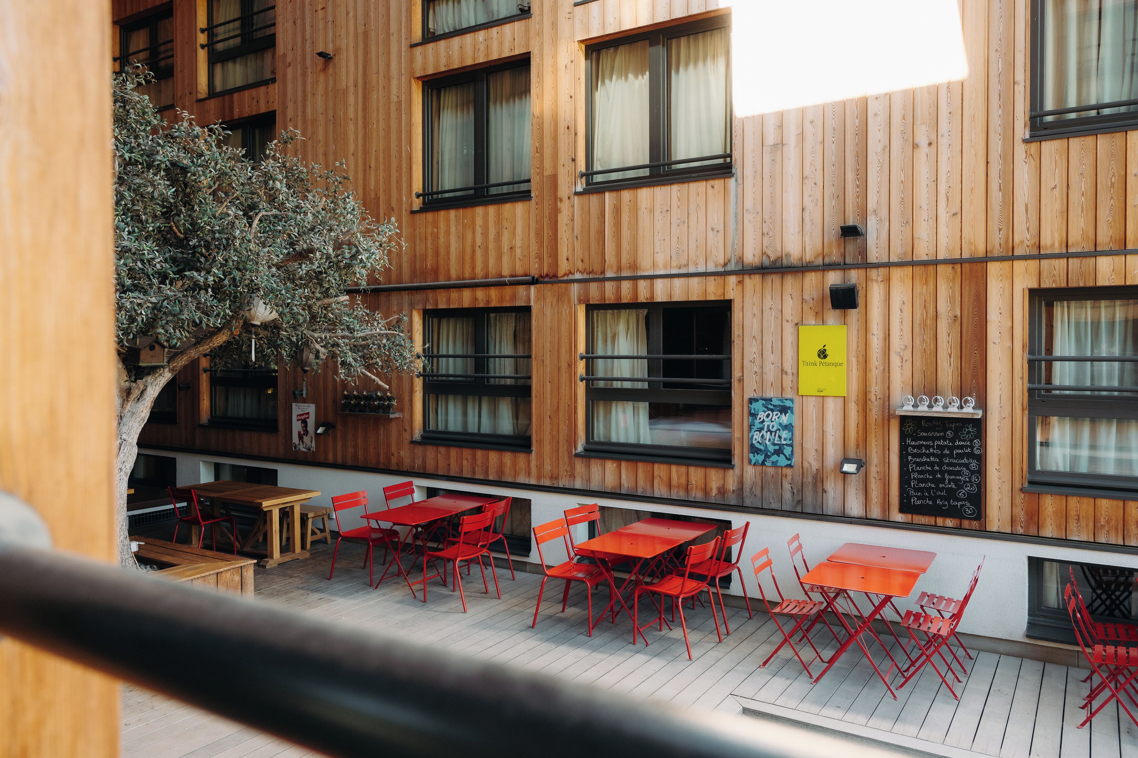 An outdoor seating area with wooden tables and red chairs, a wooden wall with windows, and a tree.