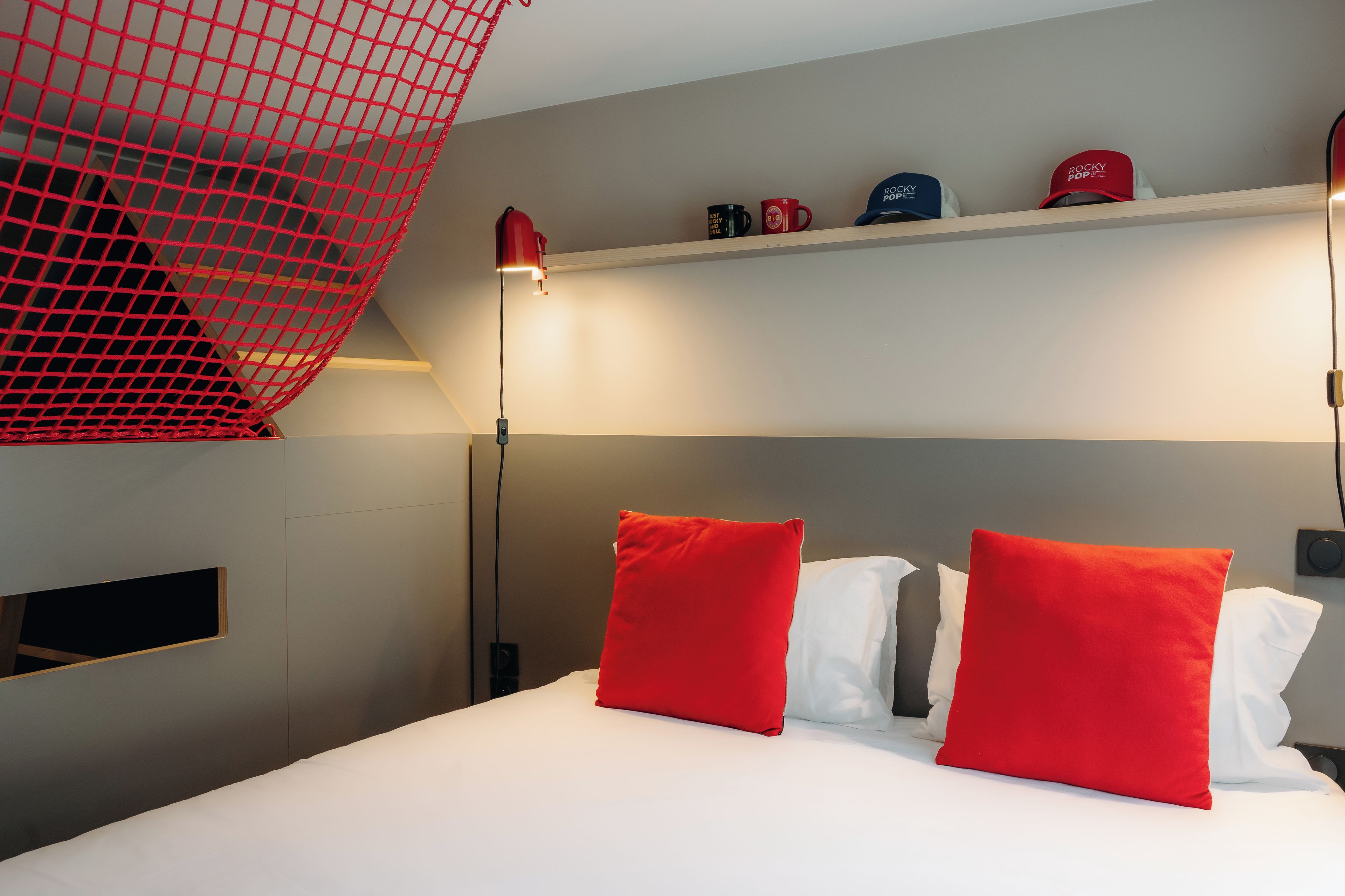 A hotel room with a bed, red and white pillows, a shelf with hats, and a red net structure.