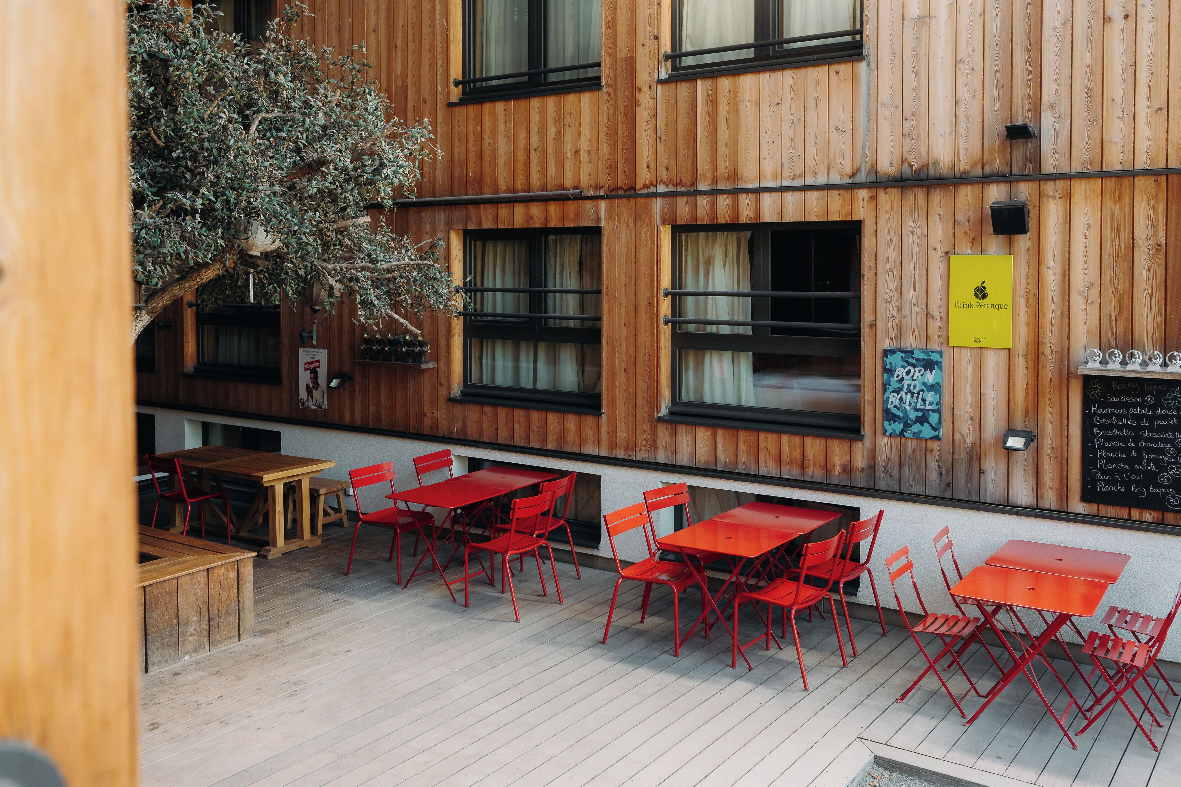 Un espace extérieur aménagé avec des tables en bois et des chaises rouges, un mur en bois doté de fenêtres et un tableau noir servant de menu.