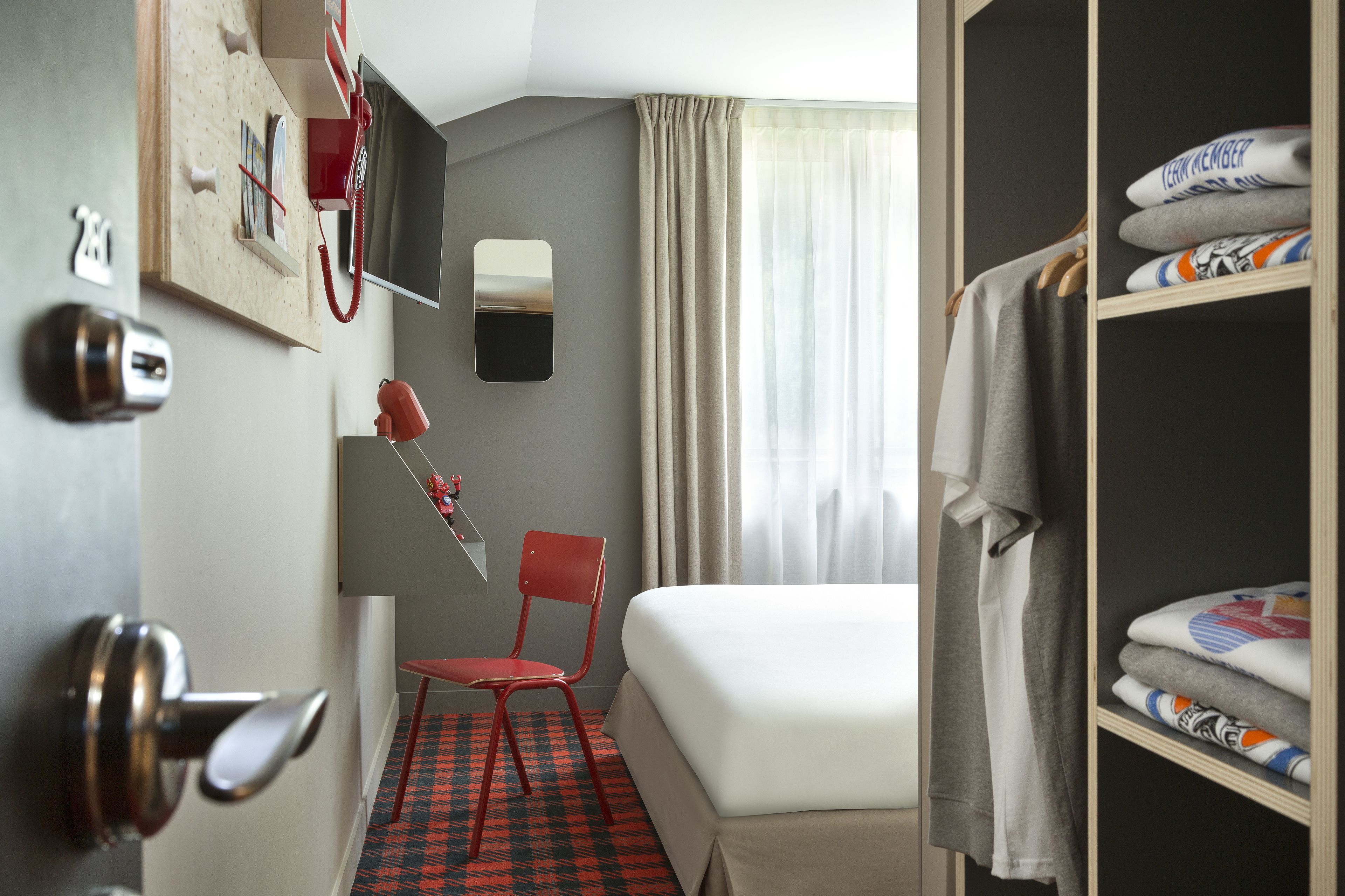 Una habitación de hotel con una cama, una silla roja, un escritorio con una computadora, un espejo y un armario.