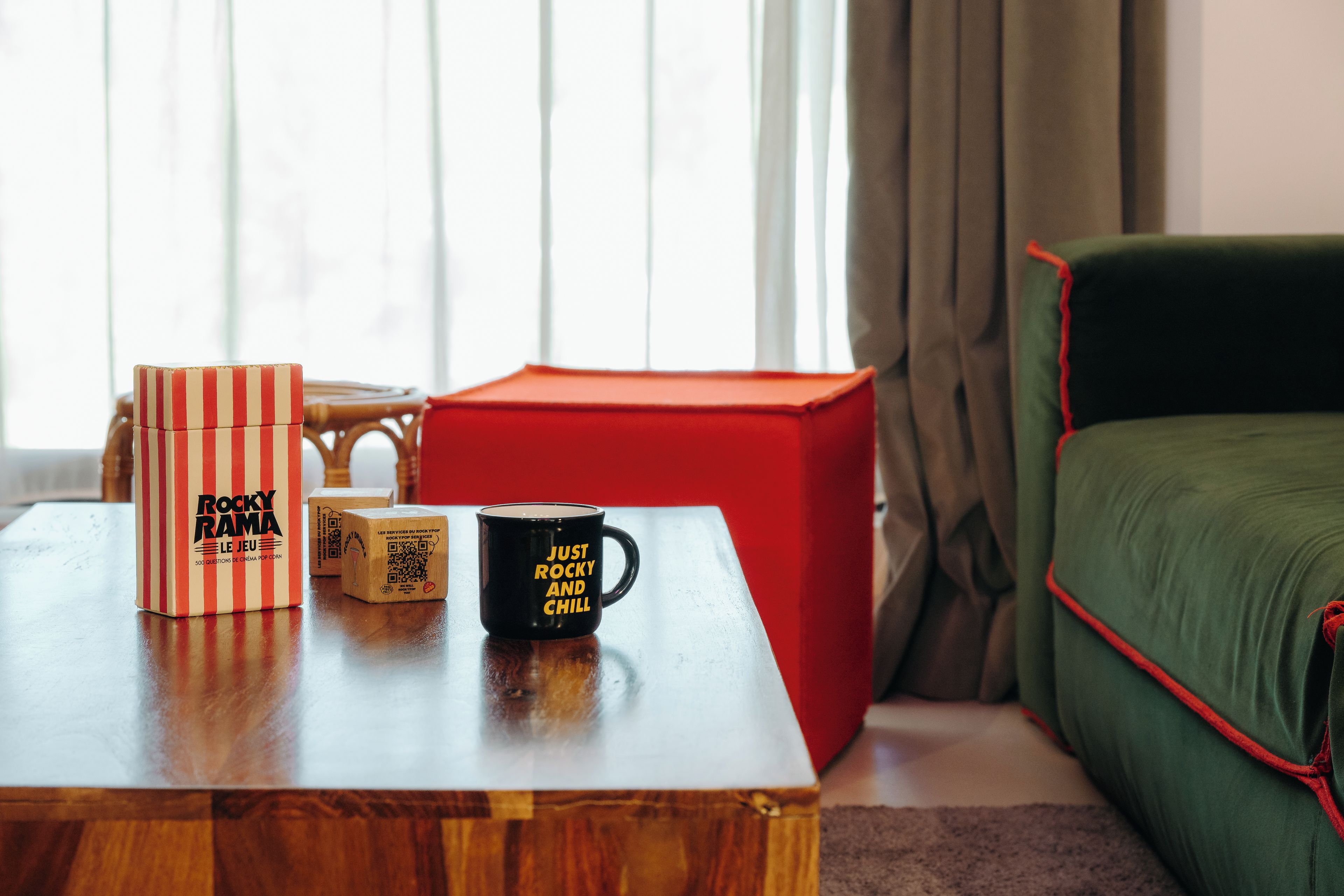 A wooden coffee table with a striped box, a black mug, and a red cube in a room with a green sofa and a window with curtains.