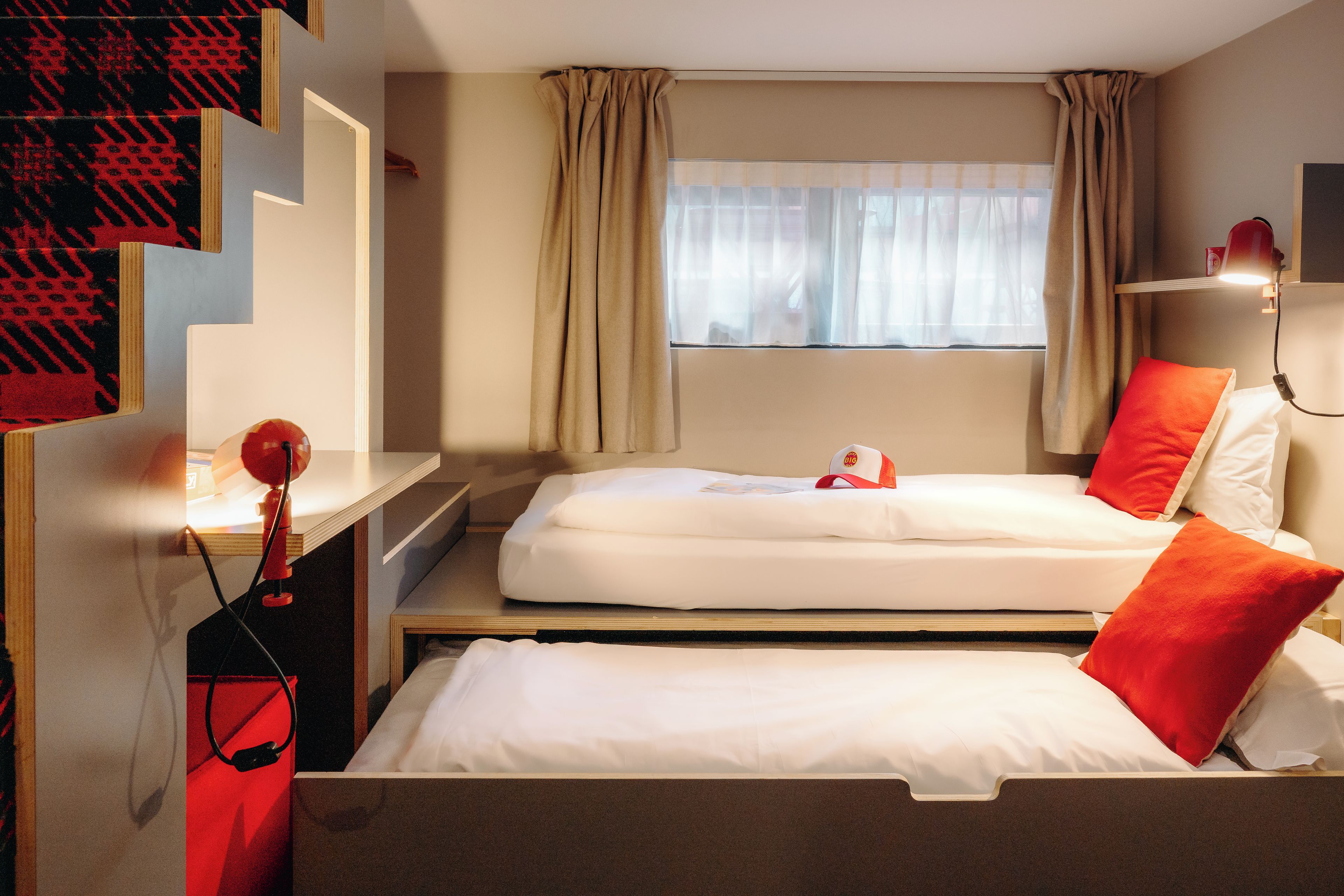 A hotel room with two beds, a desk, a chair, a window with curtains, and a wall-mounted lamp.