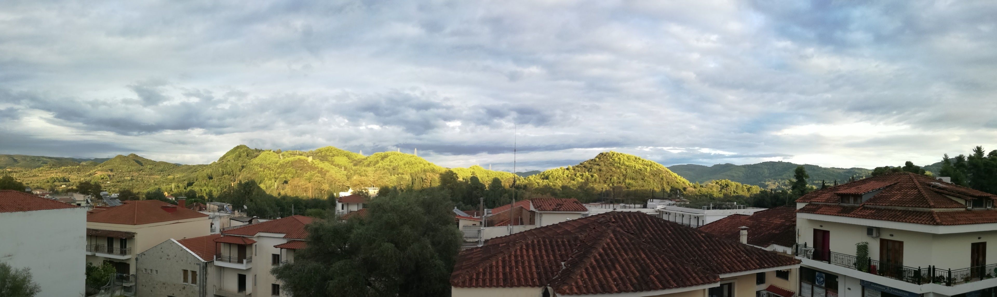 A residential area with houses featuring red-tiled roofs and white walls, surrounded by green hills and a cloudy sky.