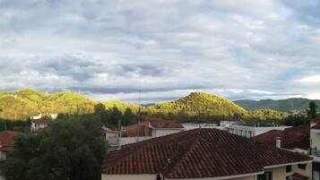 A residential area with houses featuring red-tiled roofs and white walls, surrounded by green hills and a cloudy sky.