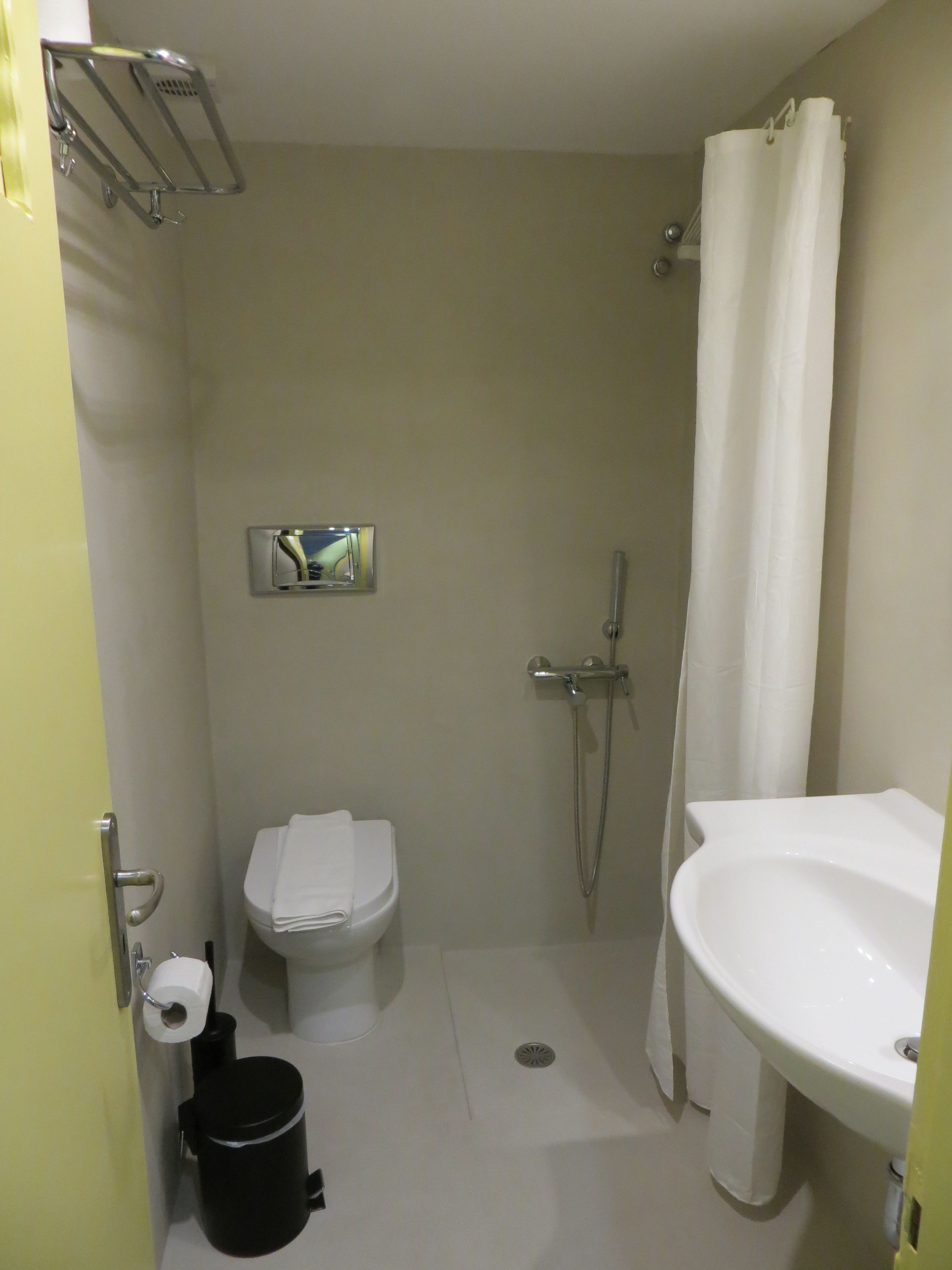 Egy kis fürdőszoba, amelyben WC, mosdókagyló, zuhanyzó és törölközőtartó található.
