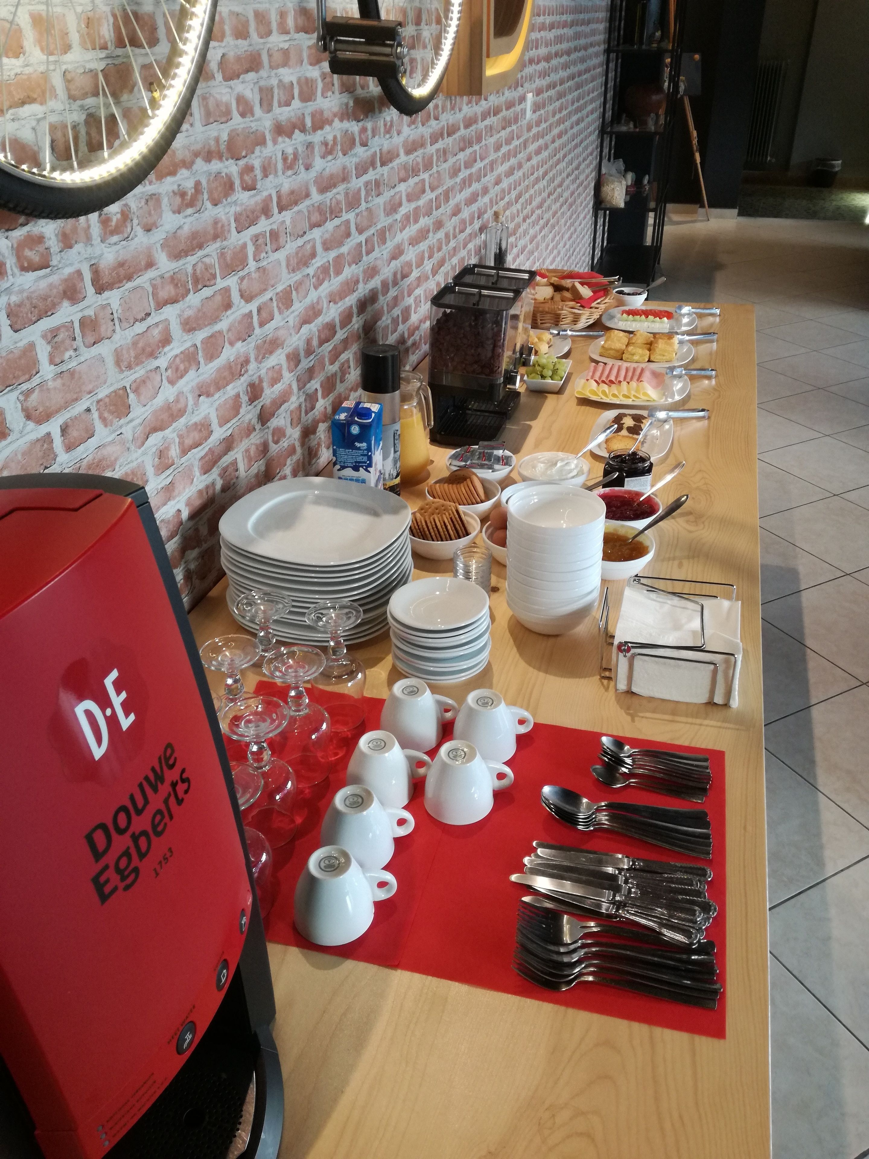 A breakfast buffet setup with plates, cups, and utensils on a table, and a red coffee machine labeled 'D.E Douwe Egberts'.