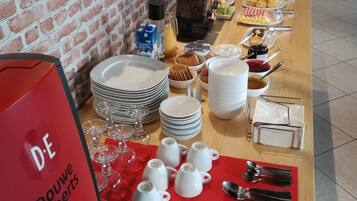 Un buffet de petit-déjeuner est installé sur une table, avec des assiettes, des tasses et des couverts, et une machine à café rouge portant l’inscription « D.E Douwe Egberts ».
