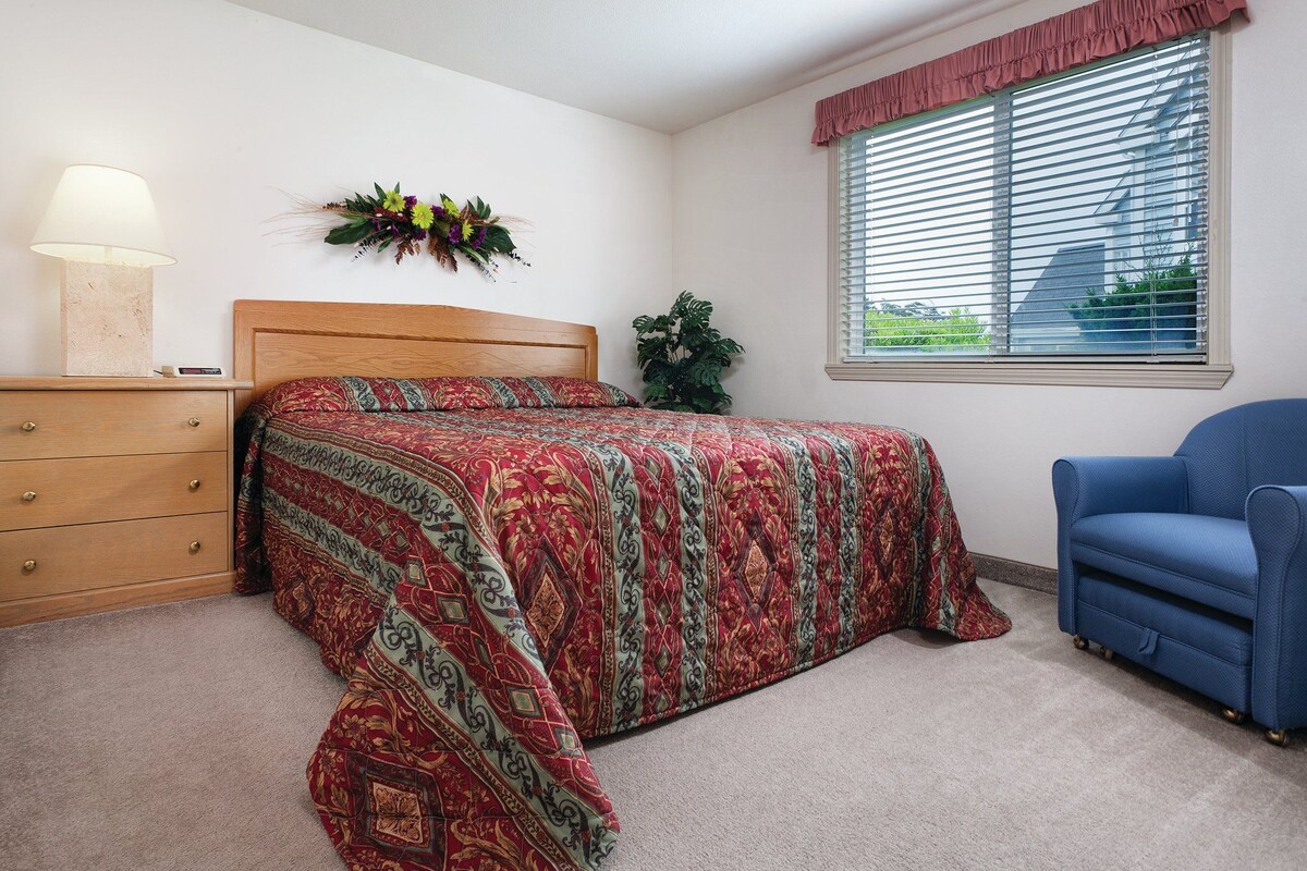 A bedroom with a bed, a dresser, a chair, a window with blinds, and a wall-mounted lamp.