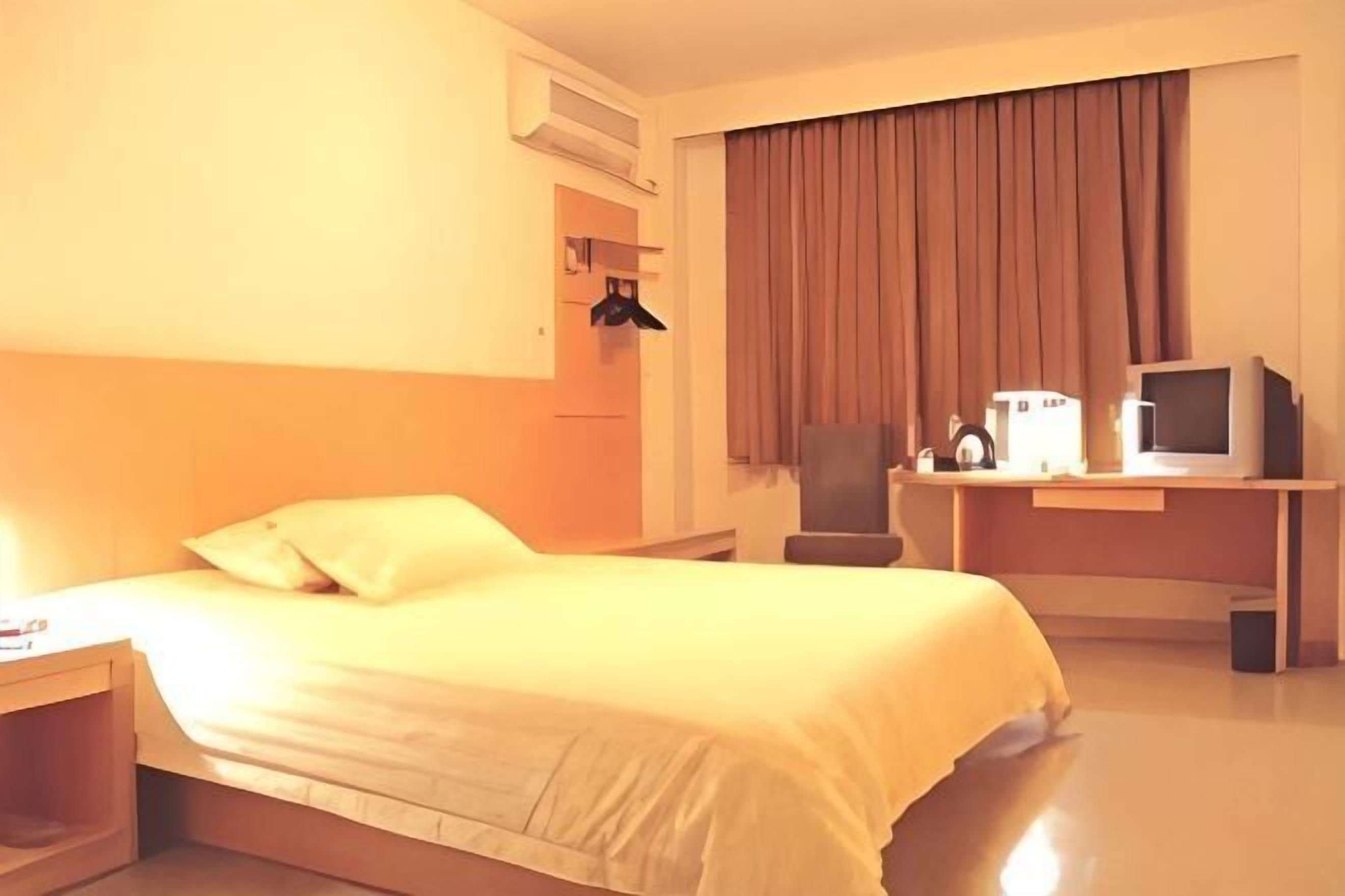 A hotel room with a bed, a desk with a computer and a chair, a TV, and a window with curtains.