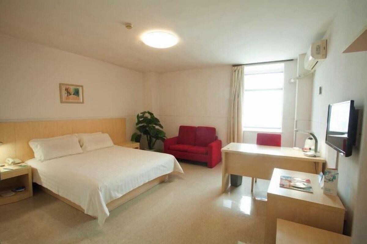 A hotel room with a bed, a desk, a red chair, a plant, and a TV.