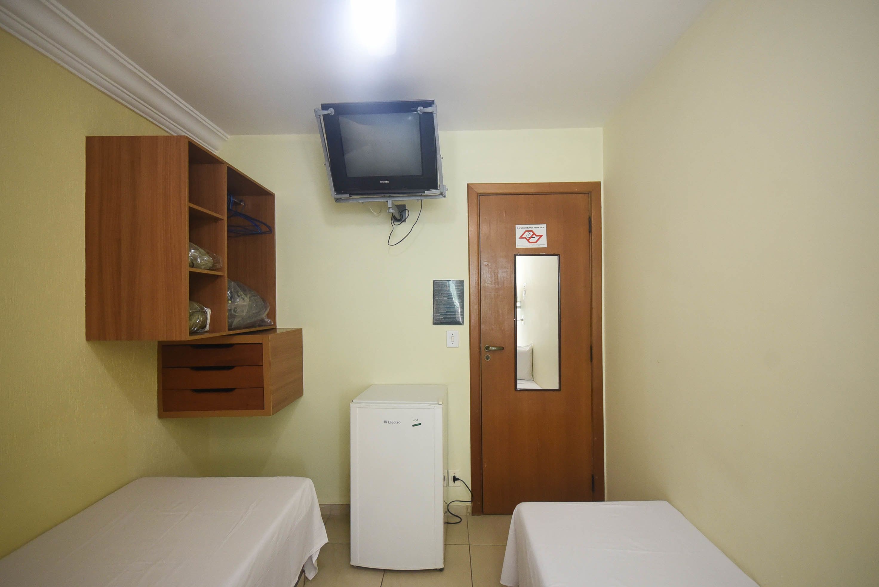 A small room with two beds, a wooden wardrobe, a white refrigerator, and a television mounted on the wall.