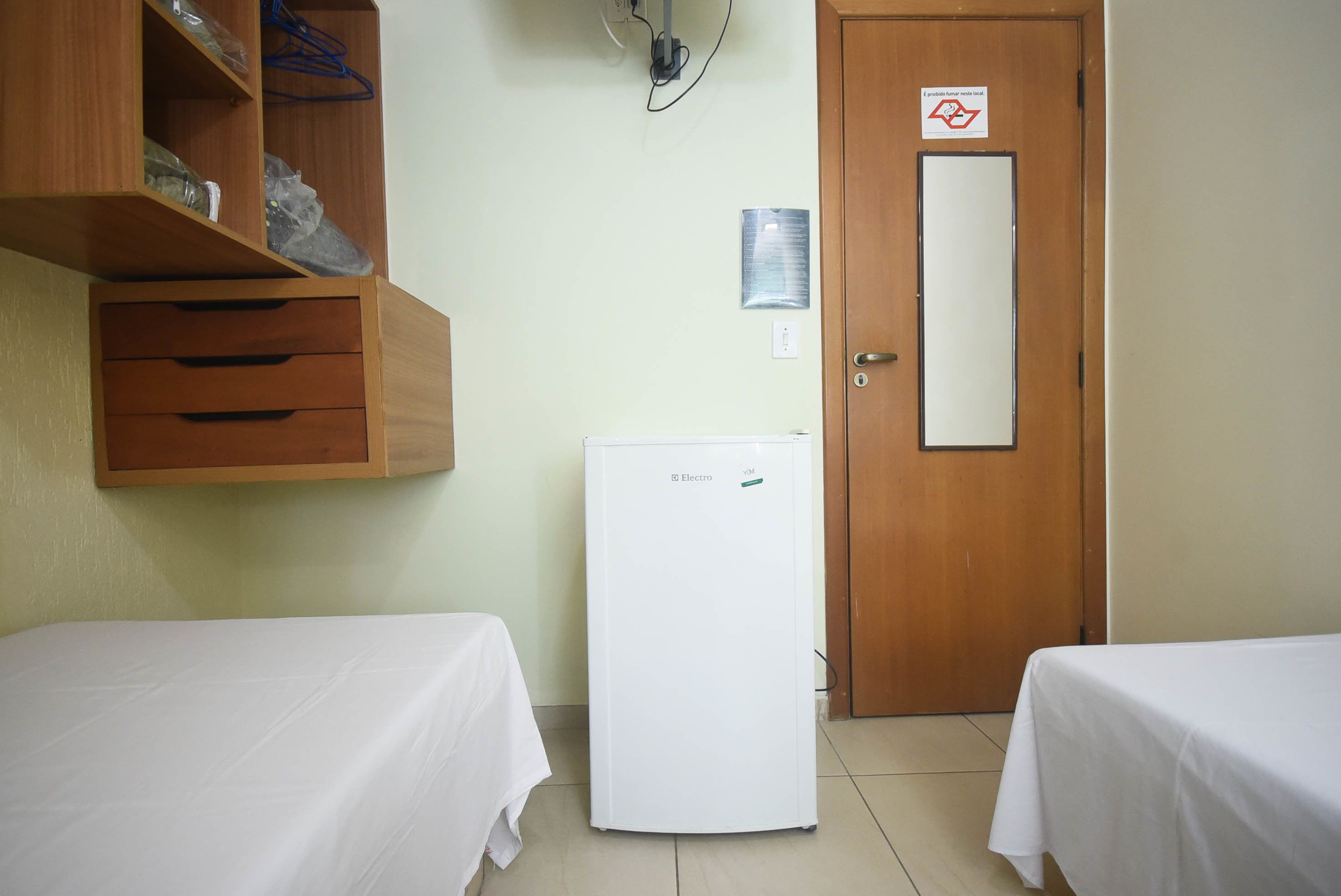 A room with two beds, a white refrigerator, wooden shelves, a door with a sign, and a wall-mounted light fixture.