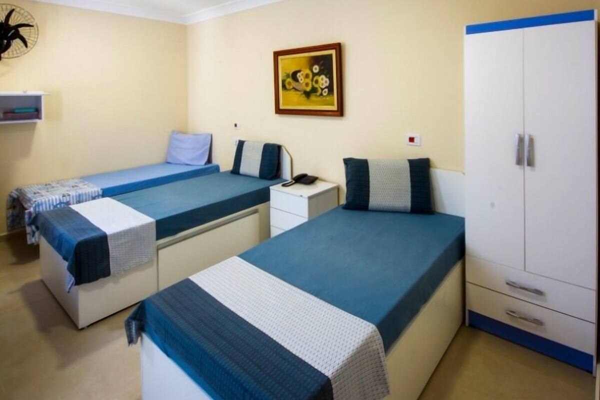 Ein Hotelzimmer mit zwei Einzelbetten, einem weißen Schrank, einem kleinen Nachttisch, einem Wandventilator und einem Gemälde.