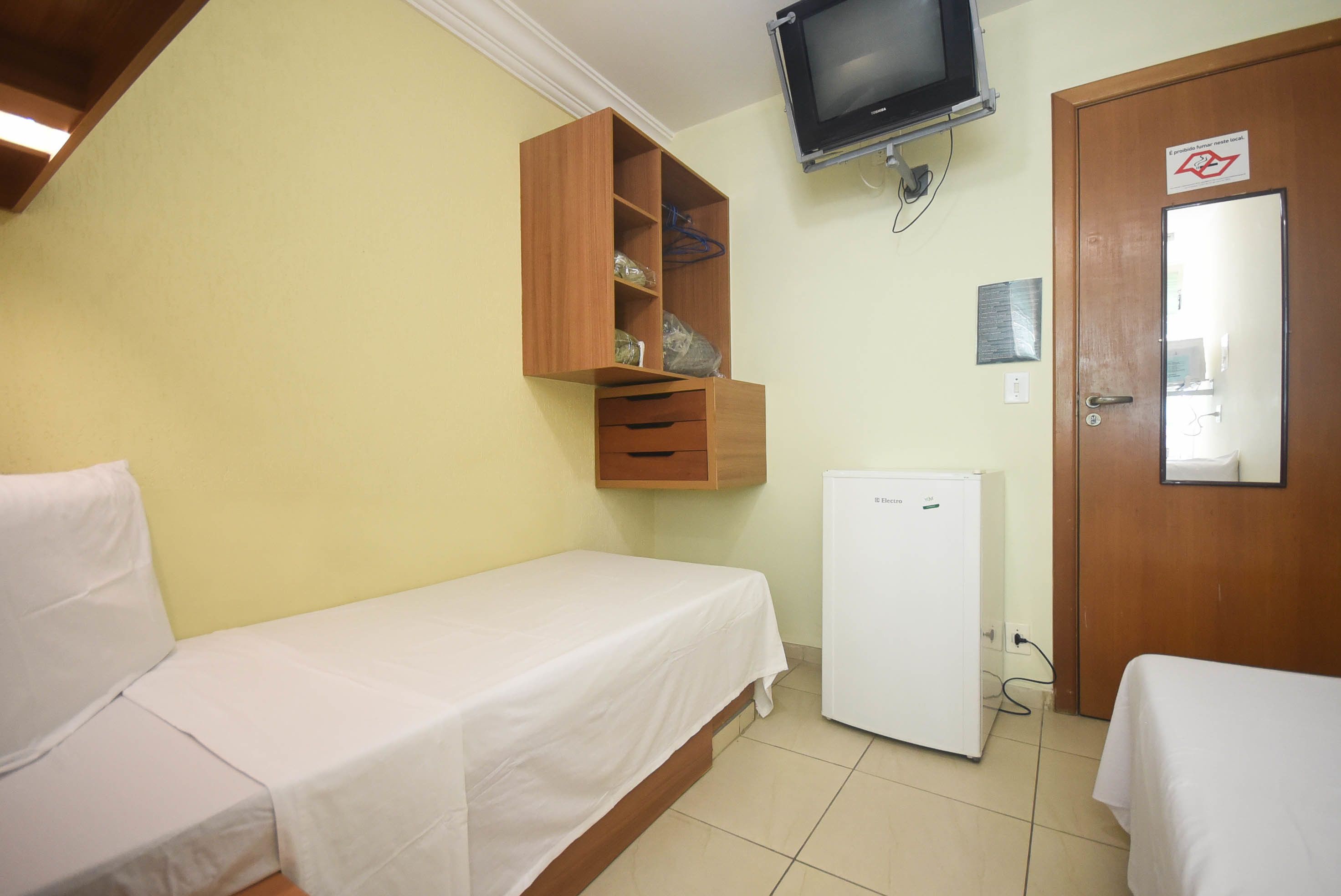 A small, single-bed room with a wooden wardrobe, a small refrigerator, and a television mounted on the wall.