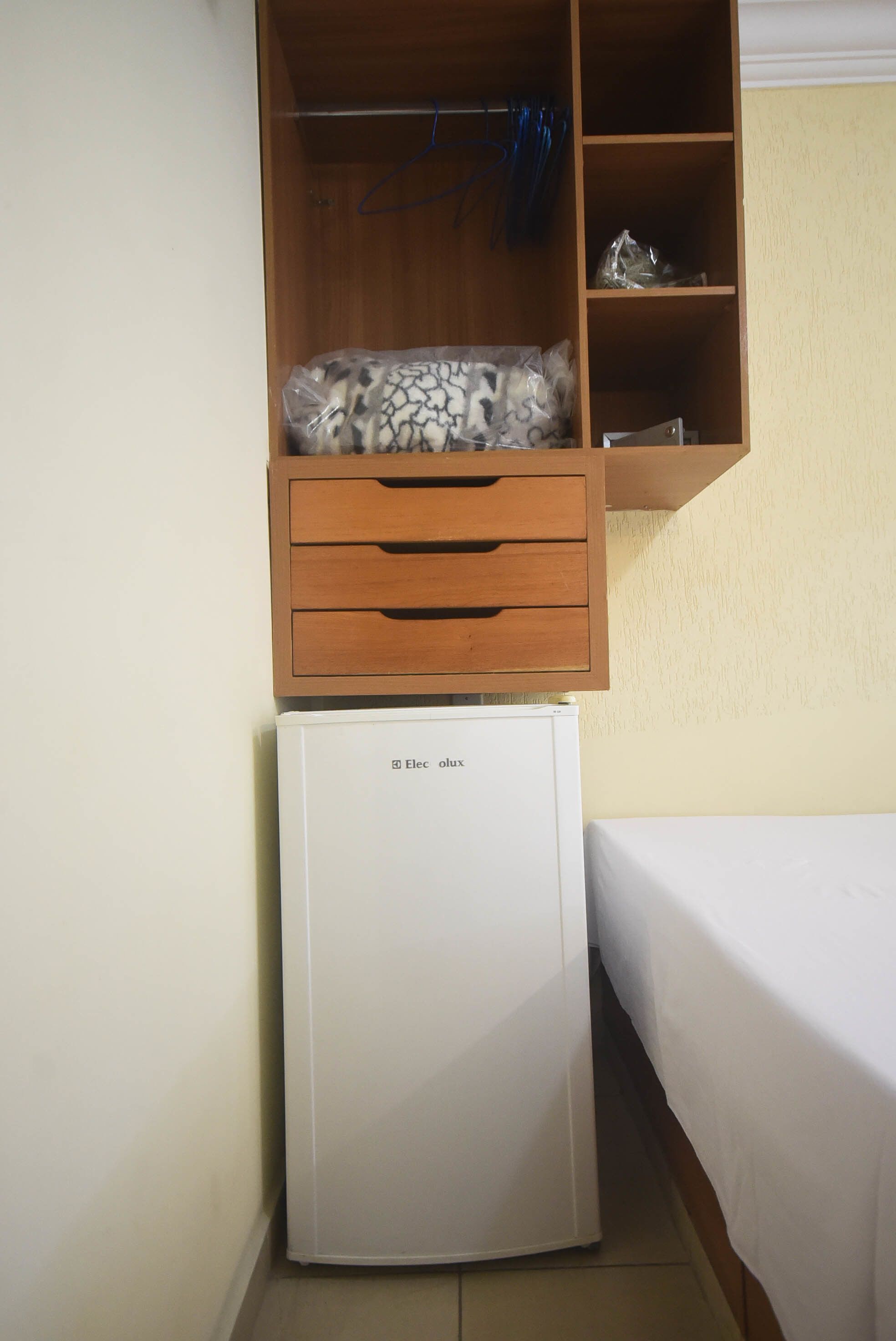 Una stanza compatta con un piccolo frigorifero bianco, un armadio in legno con abiti appesi e un letto con lenzuolo bianco.