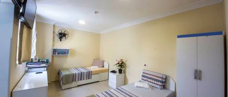 Une petite pièce bien éclairée, avec deux lits, un bureau, une armoire et un ventilateur de plafond.