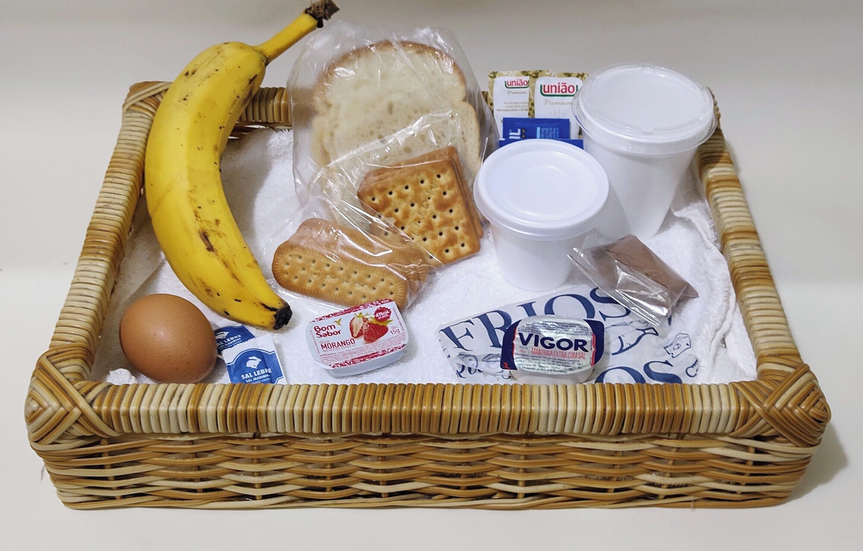 A wicker basket containing a banana, bread, crackers, a cup, a bottle, a packet of cheese, and a packet of yogurt.