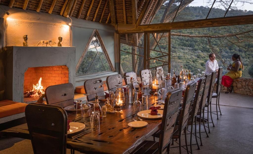 A dining area with a long wooden table set for a meal, a fireplace, and large windows offering a view of the outdoors.