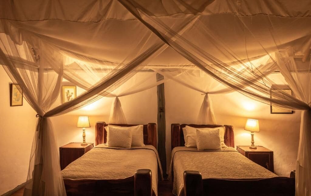A room with two beds, each with a nightstand and a lamp. The room has a canopy bed with sheer curtains.