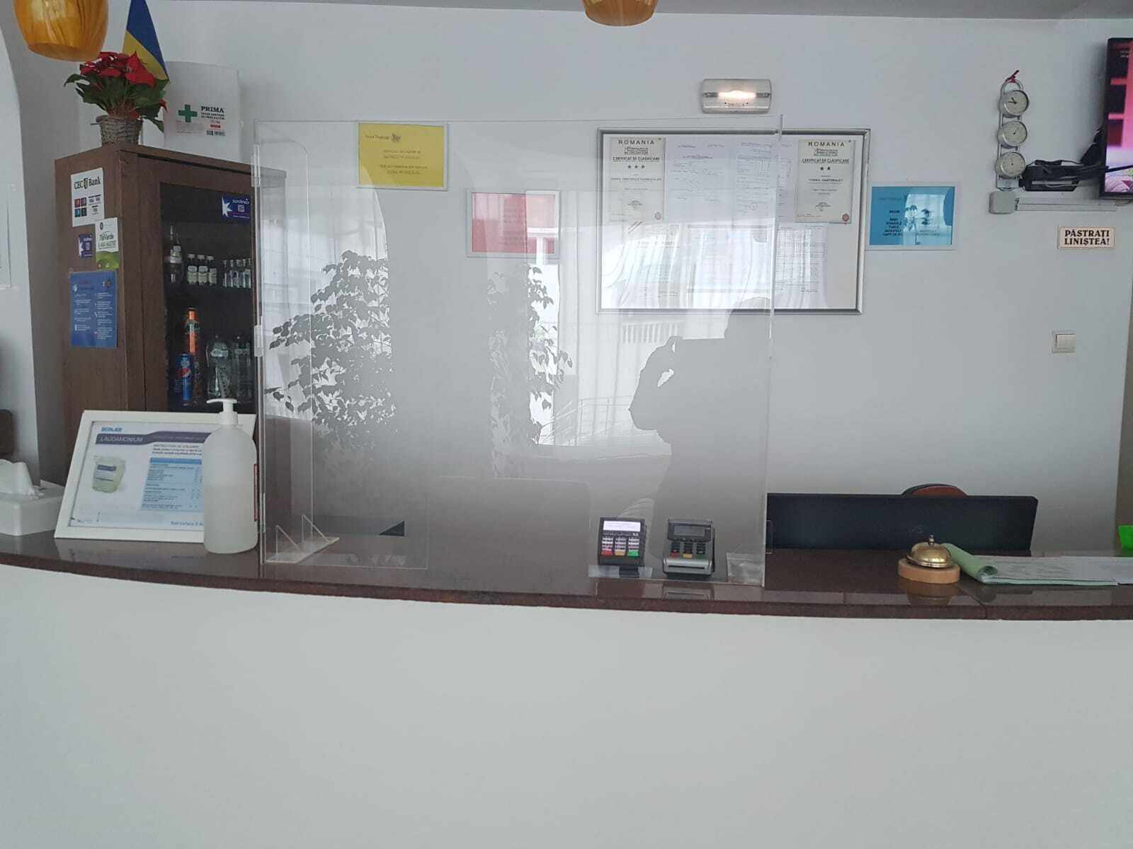 Ein Empfangsschalter mit Computer, Telefon und einer kleinen Pflanze. An der Wand befinden sich Schilder und ein Fenster mit Blick auf Bäume.
