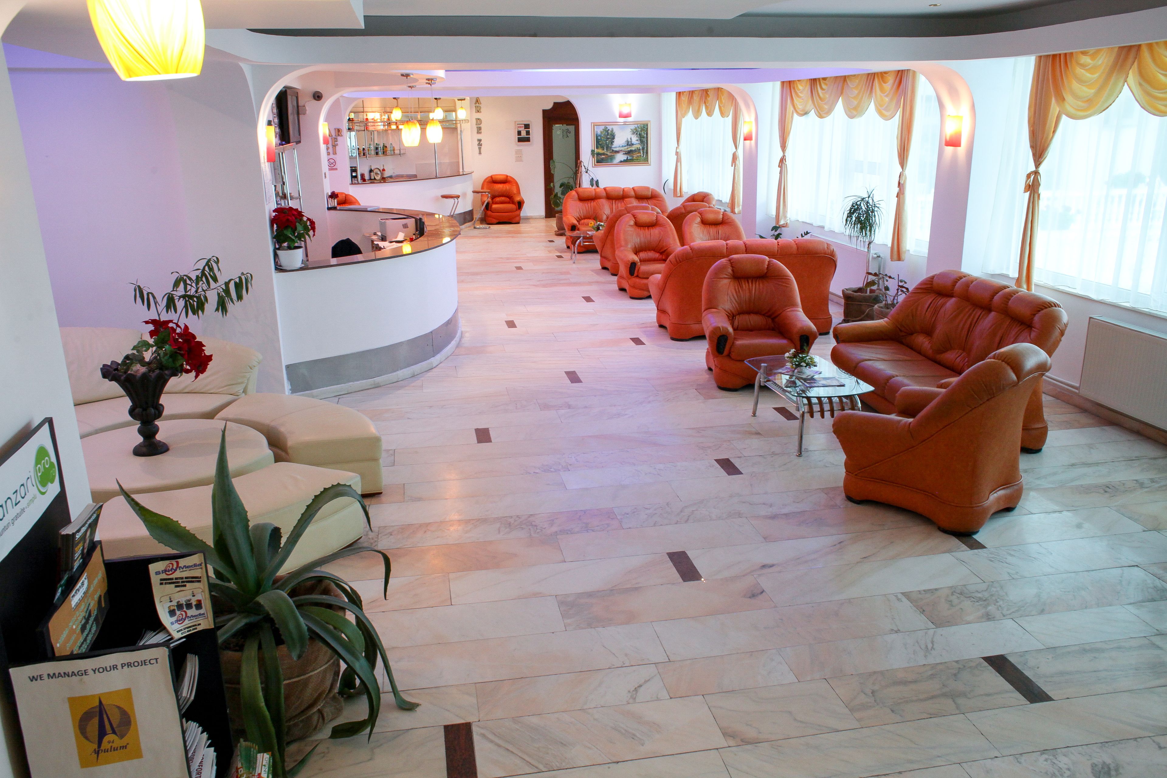 Un vestíbulo de hotel con un mostrador de recepción, varios sillones naranja, una mesa de centro y una planta grande en maceta.