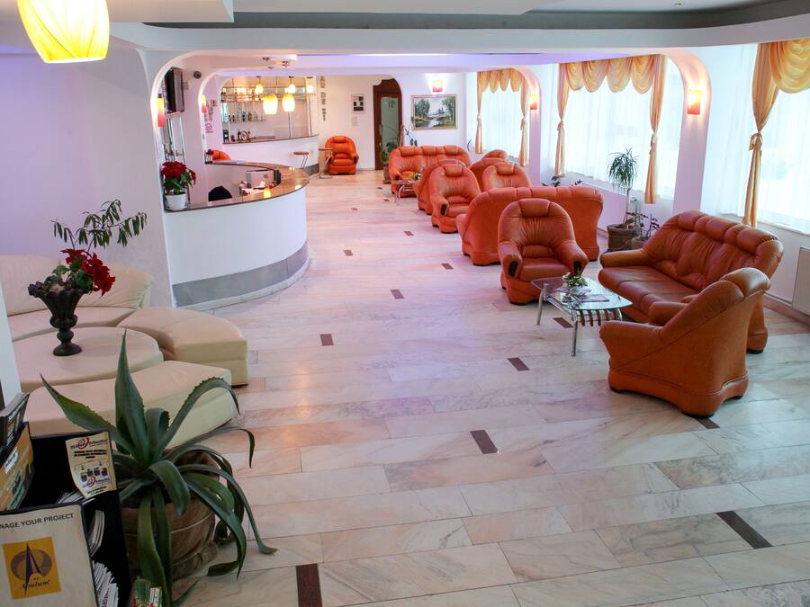 Un vestíbulo de hotel con un mostrador de recepción, varios sillones naranja, una mesa de centro y una planta grande en maceta.