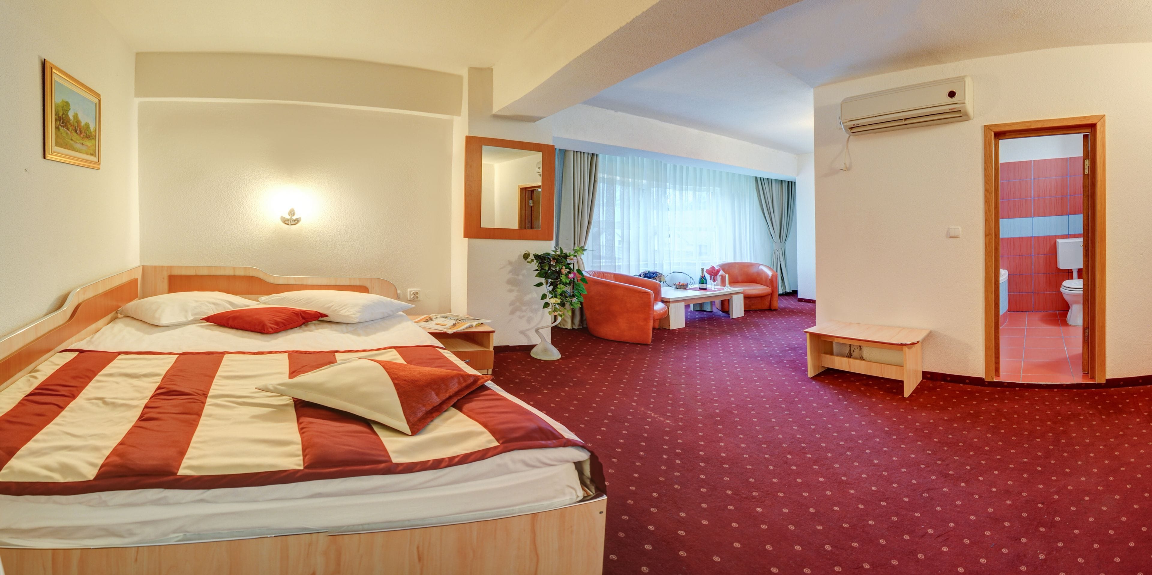 A hotel room with a large bed, a desk, a chair, and a small table. There is a mirror on the wall and a window with curtains.