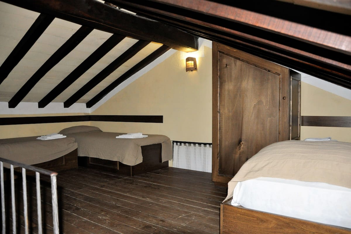 A room with a sloped ceiling, wooden beams, a bed, two smaller beds, and a wall-mounted light fixture.