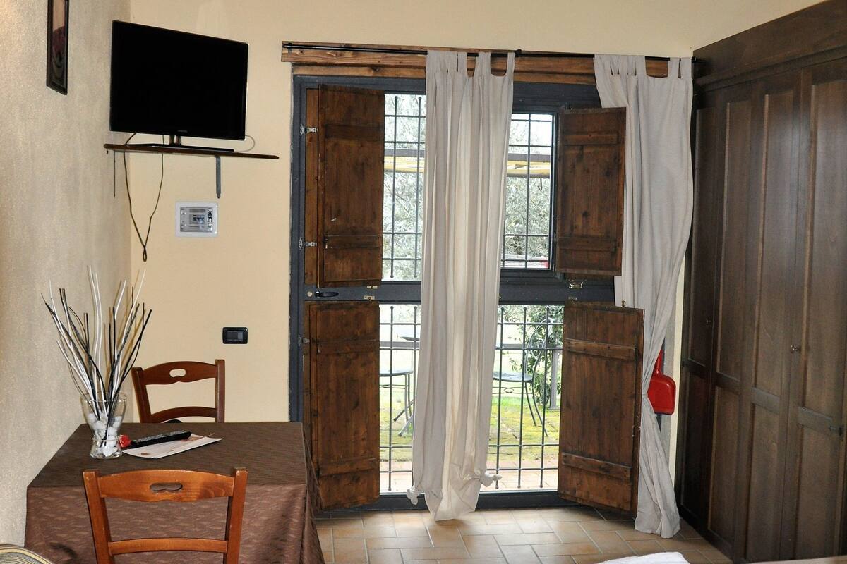 A room with a bed, a table with chairs, a TV mounted on the wall, and a window with curtains.