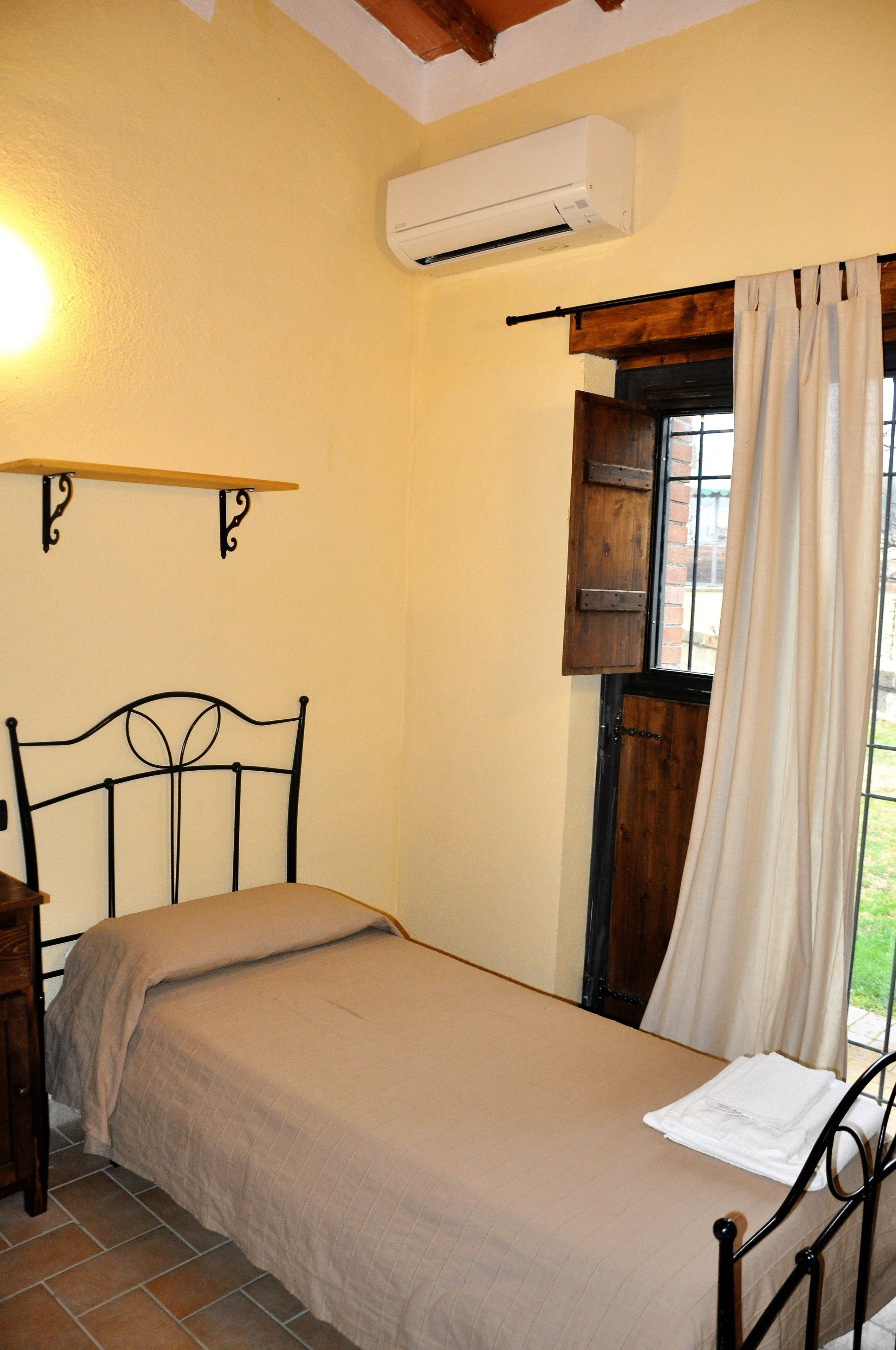 A single bed with a headboard, a bedside table, an air conditioning unit, and a window with curtains.