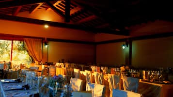 Uma sala de banquetes com mesas montadas para um evento formal, apresentando teto alto com vigas aparentes e grandes janelas que permitem entrada de luz natural.