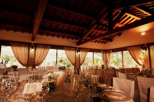 Un salón de banquetes con mesas dispuestas para un evento formal, con vigas de madera en el techo y ventanas grandes con cortinas.