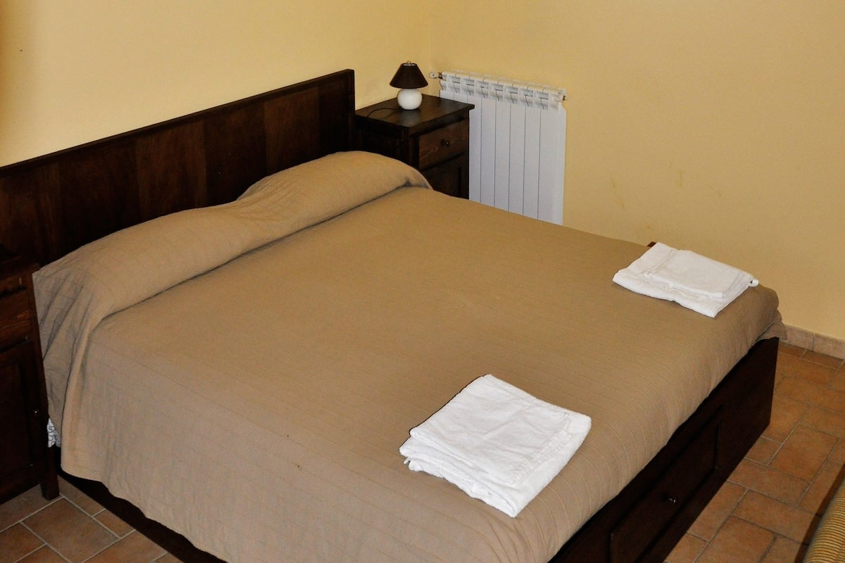 A single bed with a brown bedspread and two white towels, a wooden headboard, a nightstand with a lamp, and a radiator on the wall.