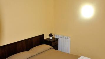 A single bed with a brown bedspread and two white towels, a wooden headboard, a nightstand with a lamp, and a radiator on the wall.