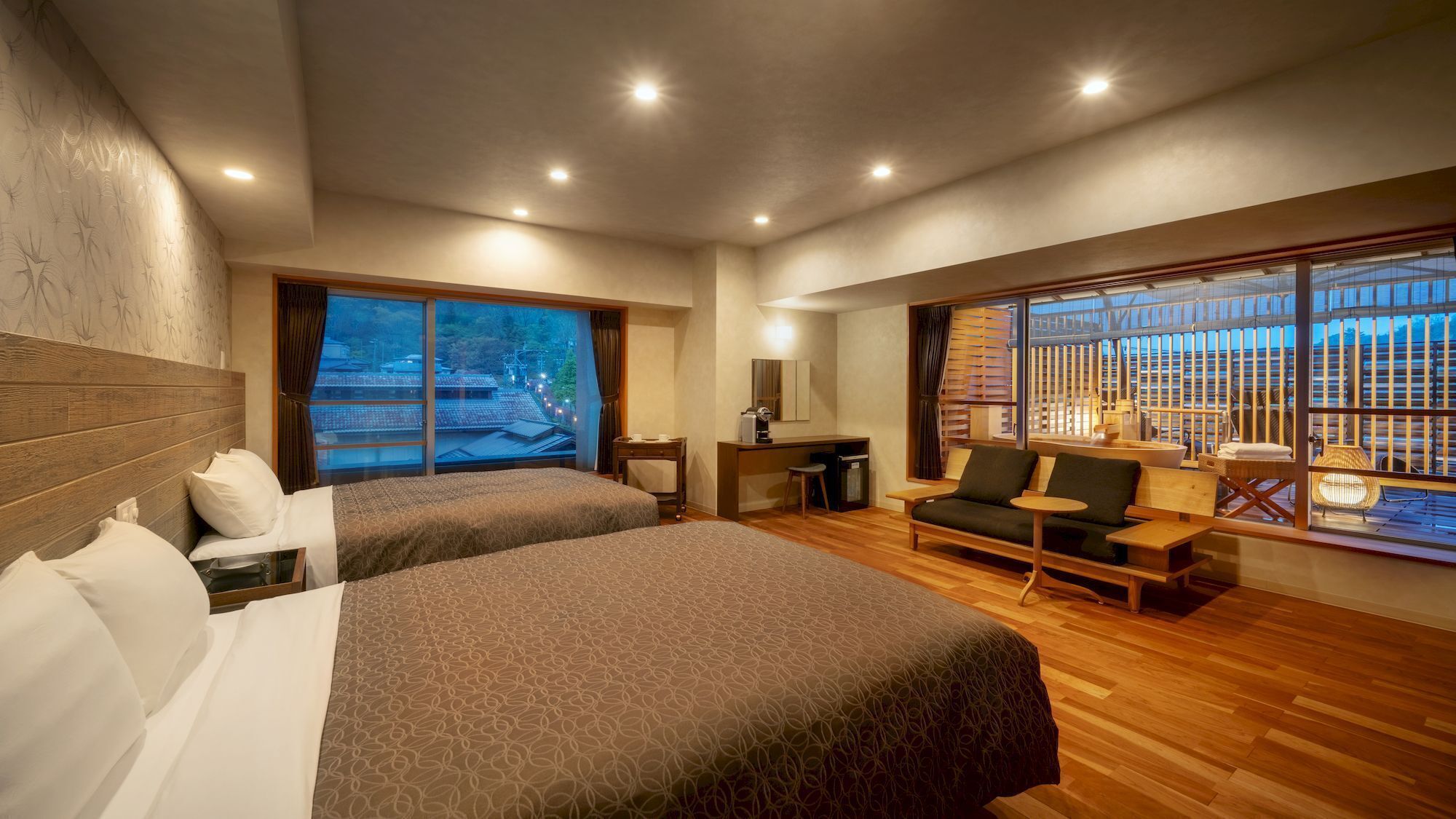 Een hotelkamer met twee bedden, een bureau, een bank en grote ramen met uitzicht op gebouwen.