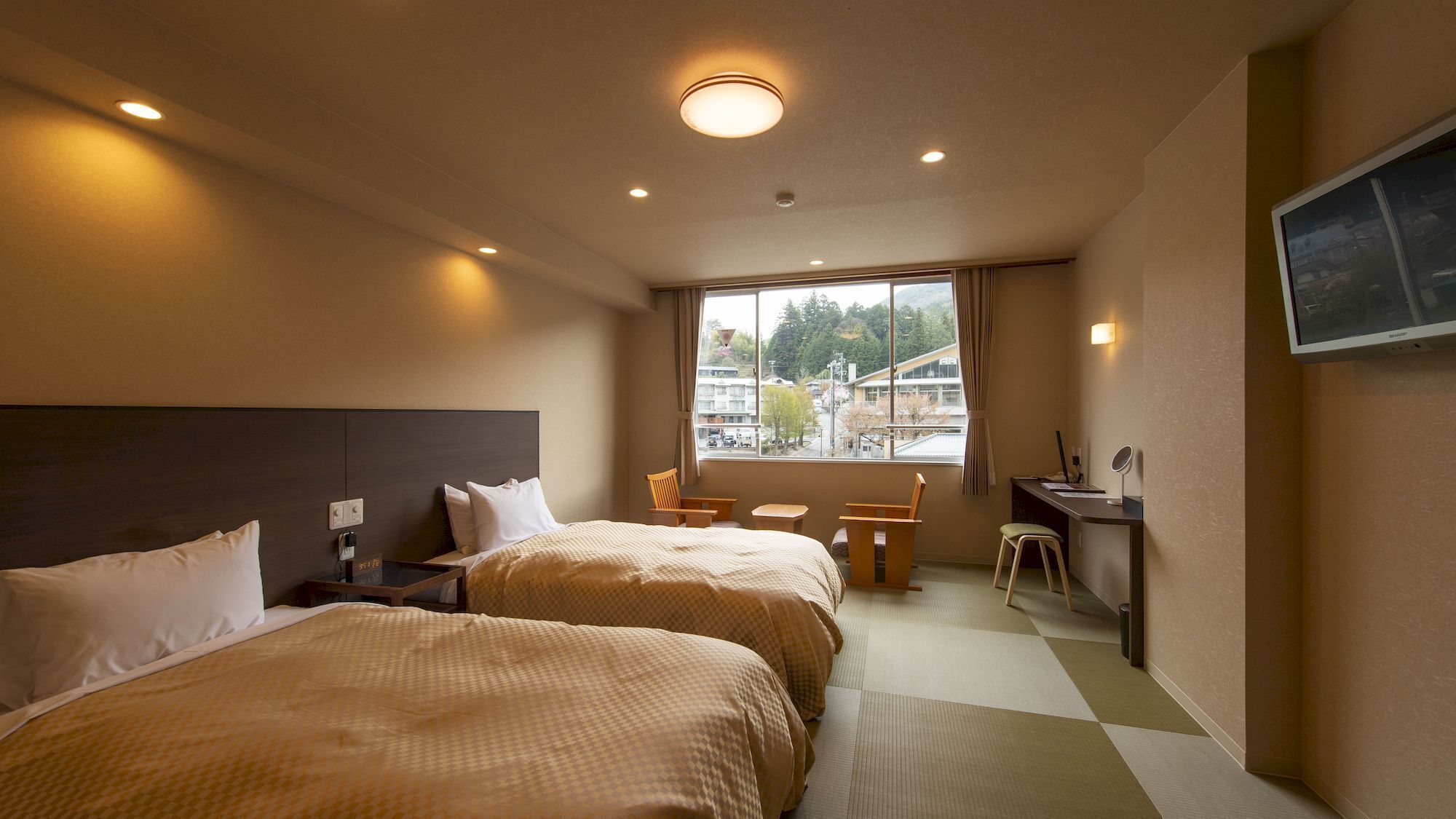Een hotelkamer met twee bedden, een bureau, een stoel, een televisie en een raam met uitzicht op gebouwen en bomen.