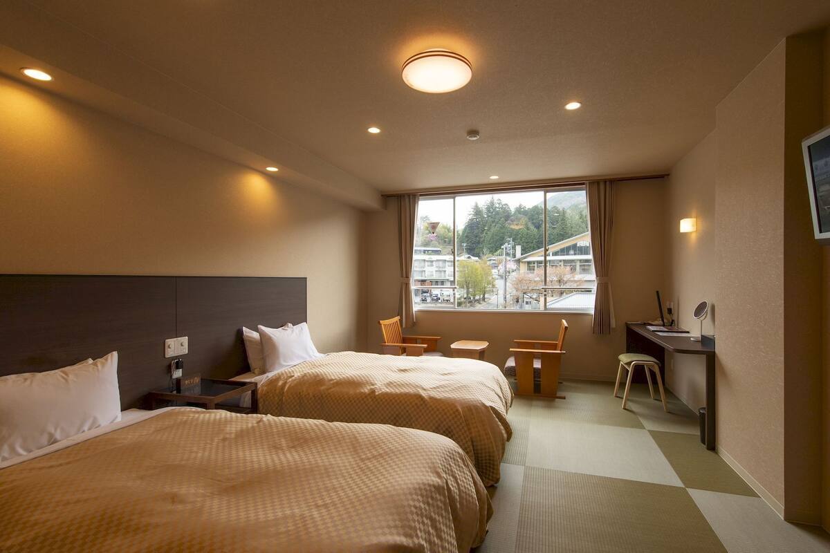 Une chambre d’hôtel avec deux lits, un bureau, une chaise, une télévision et une fenêtre donnant sur des immeubles et des arbres.