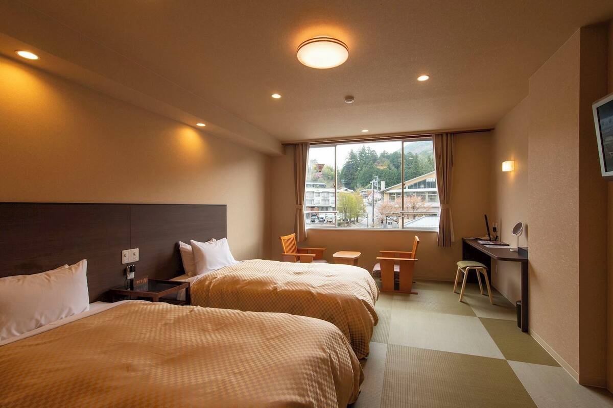 Une chambre d’hôtel avec deux lits, un bureau, une chaise, une télévision et une fenêtre donnant sur des immeubles et des arbres.