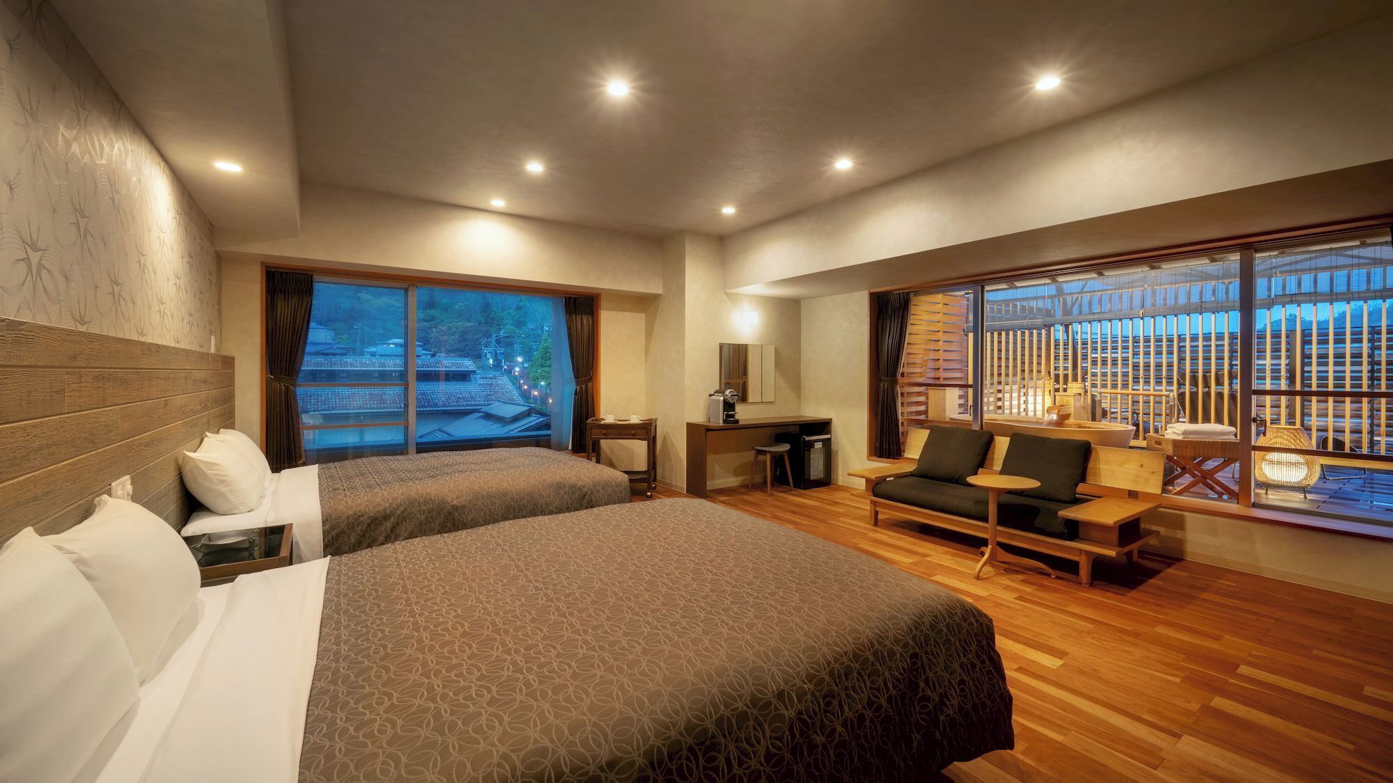 Een hotelkamer met twee bedden, een bureau, een bank en grote ramen met uitzicht op gebouwen.