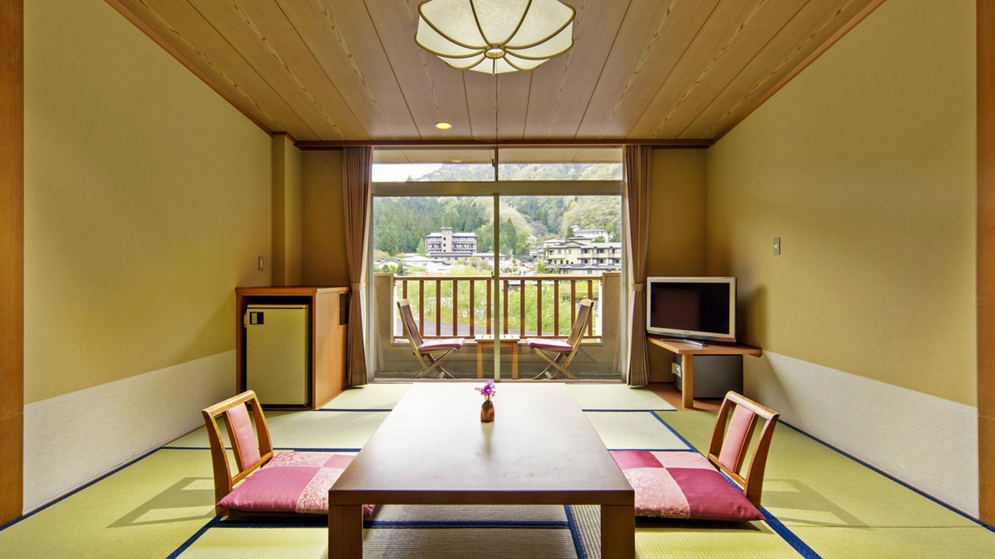 Een traditionele Japanse kamer met tatami vloerbedekking, een lage tafel, twee stoelen, een televisie en uitzicht op een bergachtig landschap.