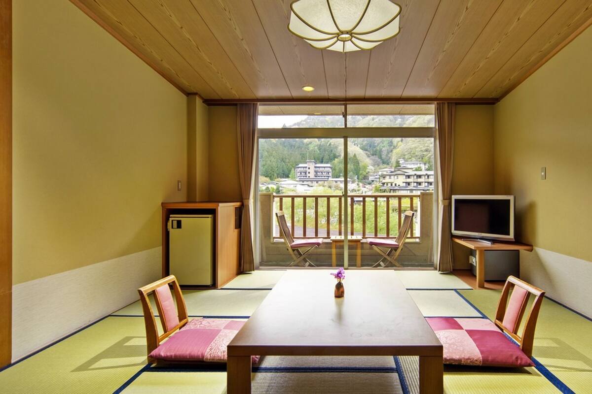 Une pièce de style japonais traditionnel, avec un sol recouvert de tatamis, une table basse, deux chaises, un téléviseur et une vue sur un paysage montagneux.