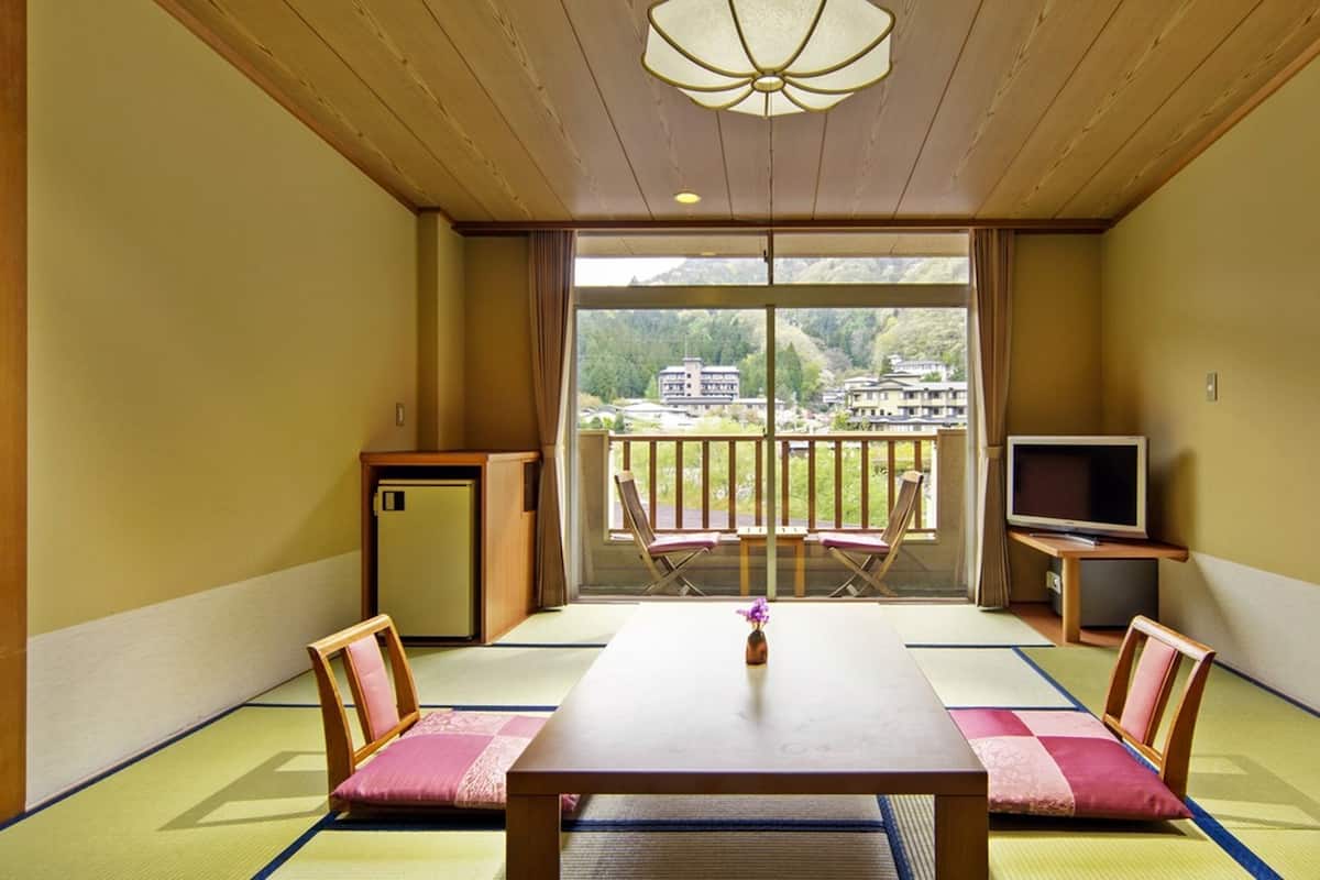 Une pièce de style japonais traditionnel, avec un sol recouvert de tatamis, une table basse, deux chaises, un téléviseur et une vue sur un paysage montagneux.