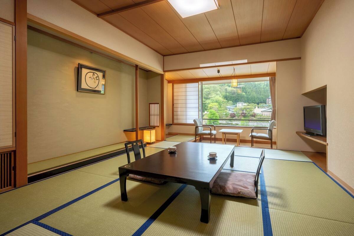 Une pièce de style japonais traditionnel, avec un sol recouvert de tatamis, une table basse, des chaises, un téléviseur et une vue sur la verdure à l’extérieur.