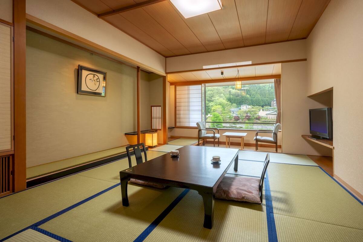 Une pièce de style japonais traditionnel, avec un sol recouvert de tatamis, une table basse, des chaises, un téléviseur et une vue sur la verdure à l’extérieur.