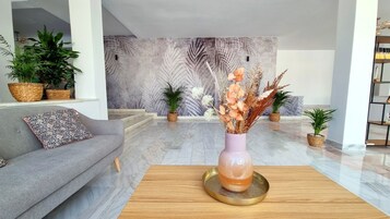 A modern living room with a grey sofa, a wooden coffee table, and a vase with dried flowers. There are potted plants on the floor and a large wall mural in the background.