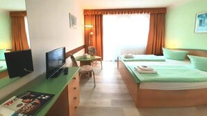 Habitación de hotel con una cama, un escritorio con televisor, una silla, una mesa con lámpara y una ventana con cortinas.